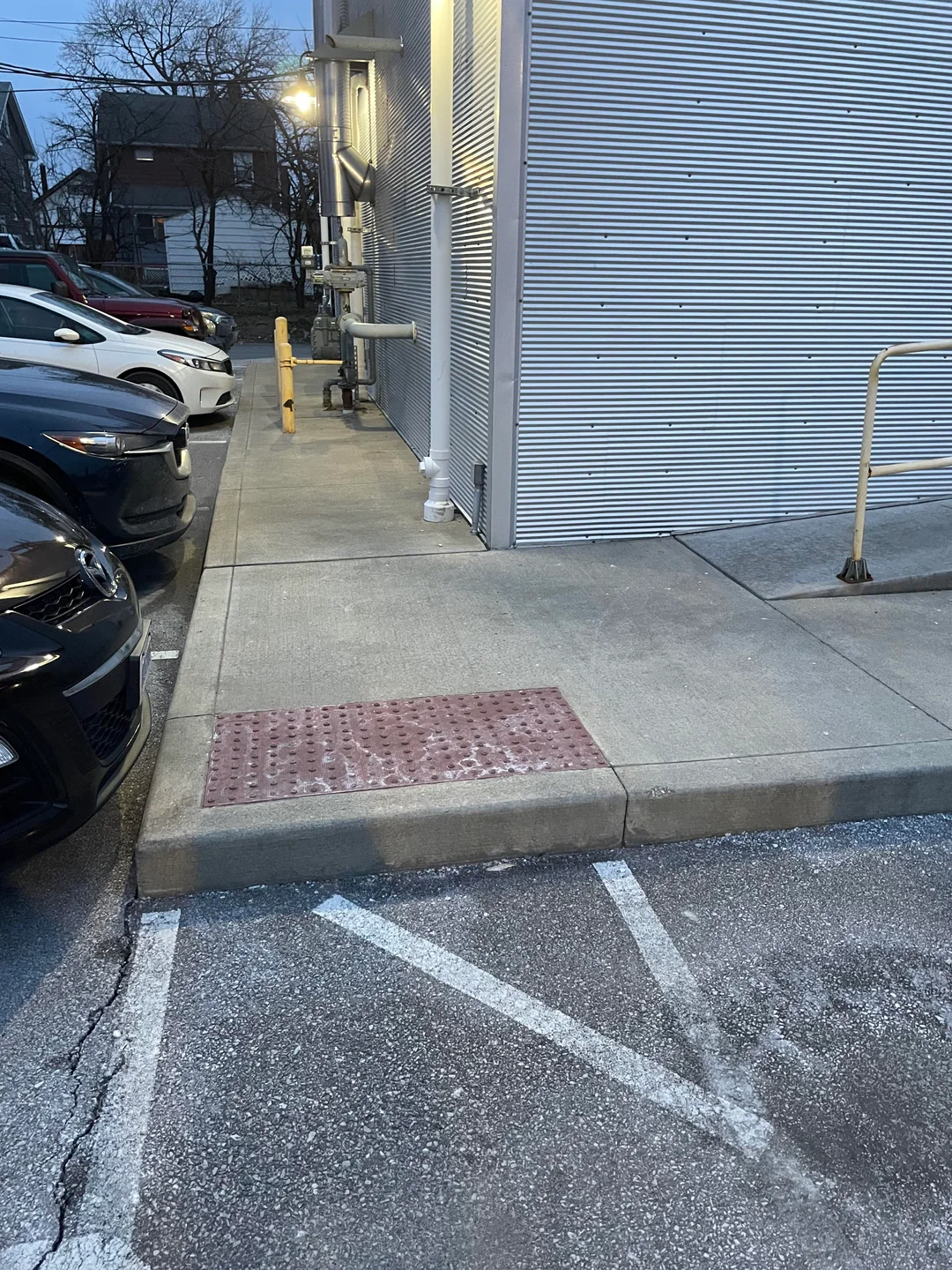 Truncated-dome panel set at the edge of a raised sidewalk beside a striped parking area, with a curb separating the sidewalk from the pavement.